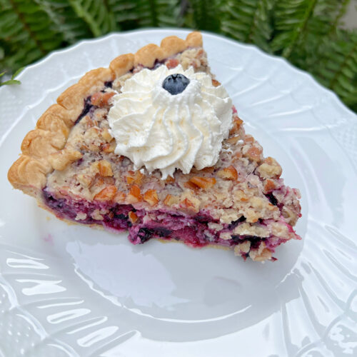 Slice of blueberry pie on a white plate, topped with whipped cream.